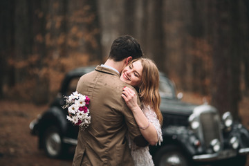 Gorgeous newlywed bride and groom posing in pine forest near retro car in their wedding day
