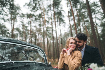 Gorgeous newlywed bride and groom posing in pine forest near retro car in their wedding day