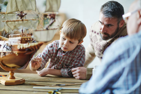 Adorable youngster painting toy ship with his father and grandfather near by