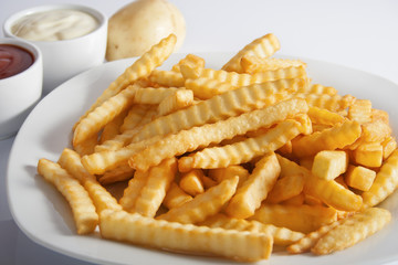 Portion of French fries (Crinkle-cut) deep fried, served on a white plate next to white bowls with mayonnaise and ketchup and fresh potato.
