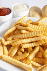 Portion of French fries (Crinkle-cut) deep fried, served on a white plate next to white bowls with mayonnaise and ketchup and fresh potato.