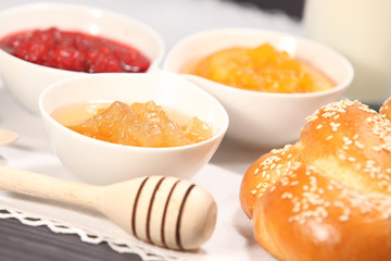 White bowls with homemade jams next to a glass of milk and sweet baked bun are on linen tablecloth on a rustic table