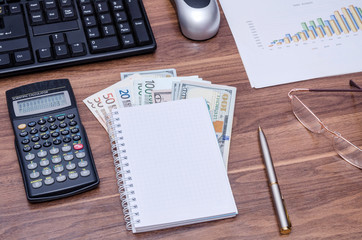 Business background, view from above on a wooden table - empty notebook and pen, dollar bills, graph, glasses, calculator, computer keyboard and mouse