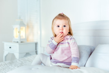 Fototapeta premium Funny amazed blond little girl sitting on bed in bedroom. White interior, big bed, bedside table and night lamp. Girl touches her mouth by hand