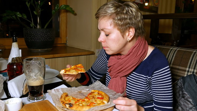 Pretty blond short hair woman eats pizza and drinks beer in cafe restaurant at the evening and talks to a friend. 4K UHD video footage.