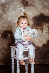 Cute blond little girl sitting on white chair and holding her toys. Studio portrait on brown grunge background