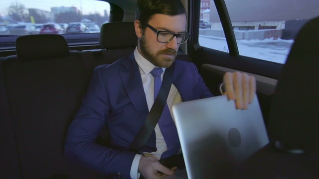 Young businessman sitting in the backseat of company car and working on his laptop