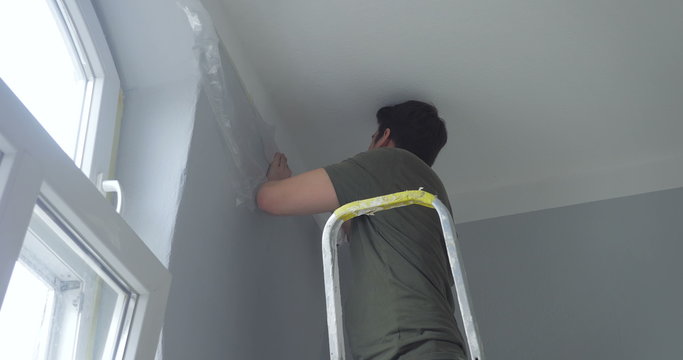 man standing on ladder and attaching ribbon tape on wall. window is on the left, it is daytime.