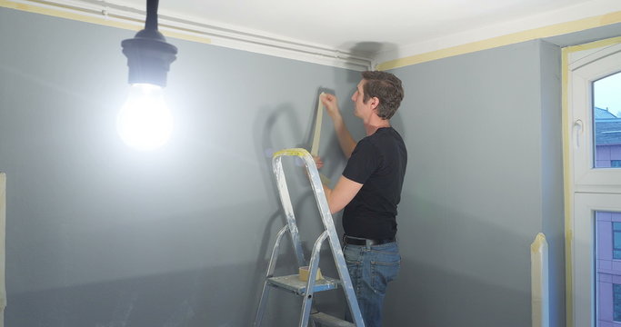 man standing on ladder and attaching ribbon tape on a wall. A light bulb hanging from ceiling as light source for the room.