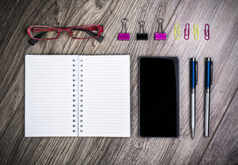 Top View Of An Office Desk With Spiral Notebook, Smart Phone, Glasses, Fountain Pen, Pen And Paperclips.