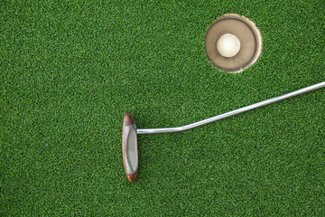 Putting golf club on green grass with golf ball in the hole - top view