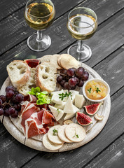Ham, cheese, grapes, figs, nuts, bread ciabatta, cracker, jam on white wooden board on bright wooden surface. Rustic style. Appetizer to wine