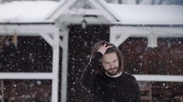 Handsome young man in black sweater standing near an old wooden house. Young bearded man in winter scenery. Snowy background. Slow motion.