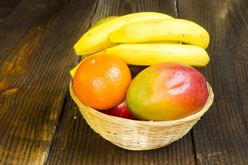 mango,klementynki i banany w wikinowym koszyczku na drewnianym tle - obrazy, fototapety, plakaty