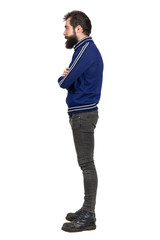 Side view of young bearded punker in blue tracksuit jacket looking away with crossed arms. Full body length portrait isolated over white studio background. 