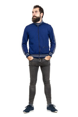 Serious bearded hipster in tracksuit jacket with hands in pockets looking away. Full body length...