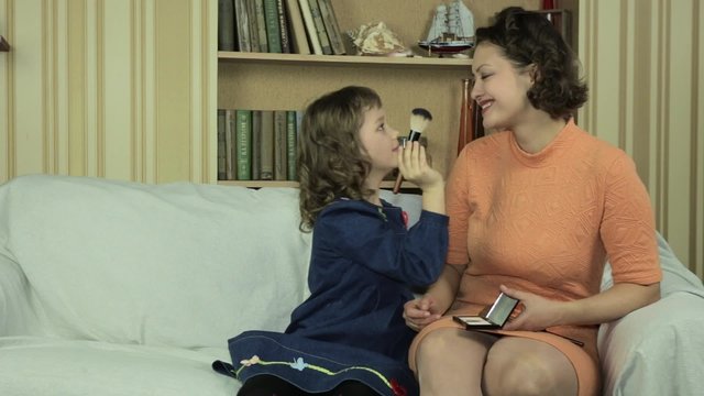 Дочка наносит макияж на лицо мамы в комнате на диване.
