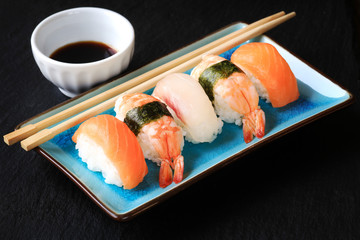 Different types of sushi on a plate with chopsticks and soy sauce on black slate background