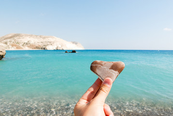 Hand holding a heart-shaped stone and Aphrodite bay in the background