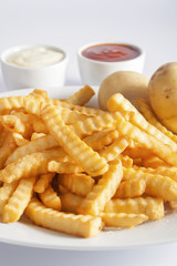 Portion of French fries (Crinkle-cut) deep fried, served on a white plate next to white bowls with mayonnaise and ketchup and fresh potato.