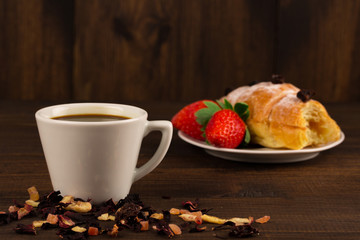 A cup of black tea with dry fruits, croissant with strawberry and wooden table background