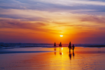 Black silhouette of happy family having horse riding adventure on sand sea beach on background of sunset sky. Active parents and people outdoor activity on tropical summer vacations with children.