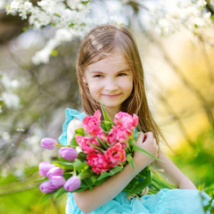 Fototapeta premium Adorable little girl holding tulips for her mother in blooming cherry garden