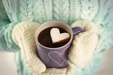 Female hands in warm mittens holding cup of hot coffee with heart marshmallow, close up