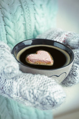 Female hands in warm mittens holding cup of hot coffee with heart marshmallow, close up