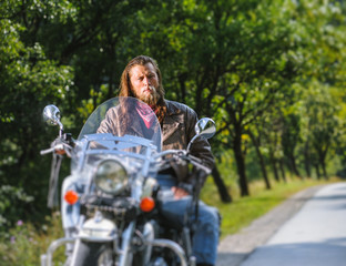 Fototapeta na wymiar Portrait of biker with long hair and beard in a leather jacket and sunglasses sitting on his motobike beside the road. Looking into the camera. Tilt shift lens blur effect