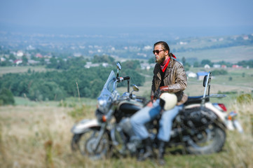 Brutal biker with beard wearing leather jacket and sunglasses sitting on his motorcycle on a sunny day and looking to the distance, holding helmet. Horizontal picture. Tilt shift lens blur effect