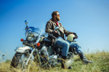 Fototapeta premium Portrait of a young man with beard sitting on his cruiser motorcycle and relaxing after driving. Man is wearing leather jacket and blue jeans. Low point of view. Tilt shift lens blur effect