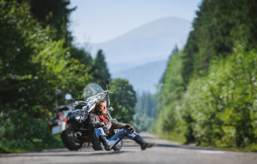 Fototapeta na wymiar Handsome man with beard and long hair sitting next to a traveler motorcycle on an open road. Guy is wearing leather jacket and blue jeans. Sunny summer day in the mountains. Tilt shift soft effect