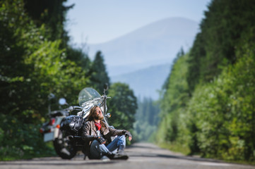 Handsome biker with beard and long hair sitting next to a traveler motorcycle on an open road looking to the sun. Guy is wearing leather jacket and blue jeans. Sunny summer day. Tilt shift soft effect