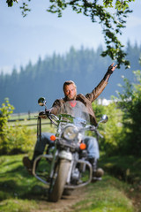 Fototapeta na wymiar young man with beard driving his cruiser motorcycle in the forest and giving the devil horns gesture. Man is wearing leather jacket and blue jeans. Tilt shift lens blur effect
