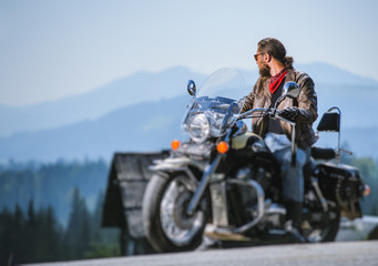 Brutal biker with beard in sunglasses, blue jeans and a leather jacket sitting on the travel motorbike and looking to the mountains. Sunny day in the mountains. Tilt shift soft effect