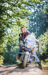 Fototapeta na wymiar Serious biker with beard driving his cruiser motorcycle in the forest. Man is wearing leather jacket and blue jeans. Tilt shift lens blur effect