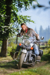 Fototapeta na wymiar Serious man with beard driving his cruiser motorcycle in the forest. Man is wearing leather jacket and blue jeans. Tilt shift lens blur effect