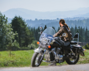 Fototapeta na wymiar Long-haired biker in sunglasses jeans and a leather jacket sitting on a black custom motorcycle and looking into distance. Sunny day in the mountains. View from the back. Tilt blur effect