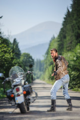Fototapeta premium Happy biker with long hair wearing leather jacket blue jeans leather boots and gloves standing near his custom made cruiser motorcycle on the open road. Tilt shift soft effect