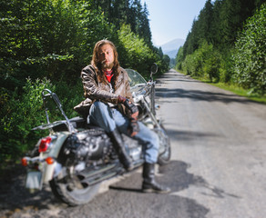 Tired biker with long hair and beard wearing a leather jacket gloves and leather boots sitting on his motorcycle on the open road. Sunny day in the mountains. Tilt shift soft effect