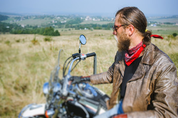 Fototapeta na wymiar Portrait of biker with long hair and beard in a leather jacket and sunglasses sitting on his bike on the grassy field. Looking into the distance. Side view. Tilt shift lens blur effect