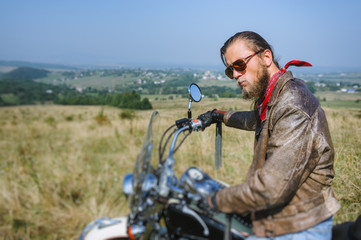 Fototapeta na wymiar Portrait of serious biker with long hair and beard in a leather jacket and sunglasses sitting on his bike on the grassy field. Looking into the distance. Side view. Tilt shift lens blur effect