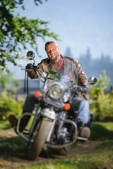 Fototapeta na wymiar young biker with beard driving his cruiser motorcycle in the forest and smiling. Man is wearing leather jacket and blue jeans. Tilt shift lens blur effect