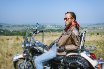 Portrait of biker with long hair and beard in a leather jacket and sunglasses sitting on his bike and relaxing on the grassy field. Looking into the distance. Side view. Tilt shift lens blur effect