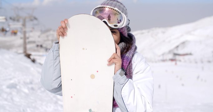 Laughing young woman wearing fashionable ski clothes and goggles posing with her snowboard on a snowy mountain at a ski resort