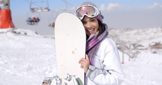 Laughing young woman wearing fashionable ski clothes and goggles posing with her snowboard on a snowy mountain at a ski resort