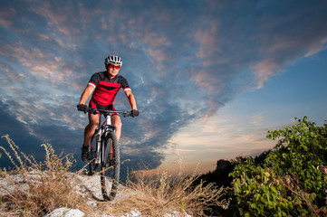 Naklejka premium Happy man on a mountain bike races downhill in the nature against blue cloudy evening sky. Cyclist is wearing red sportswear helmet gloves and red glasses. Cross country biking.