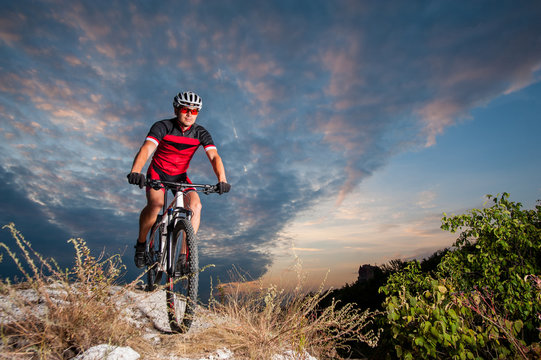 Happy man on a mountain bike races downhill in the nature against blue cloudy evening sky. Cyclist is wearing red sportswear helmet gloves and red glasses. Cross country biking.