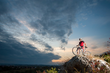 Young man athlete standing on top of a mountain with bicycle and enjoying view, with sky background.